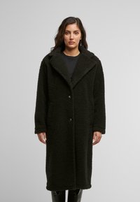 Cappotto in finto pelliccia nera con finitura testurizzata, ampio collare, chiusure frontali con bottoni e tasche laterali, progettato per una vestibilità ampia.