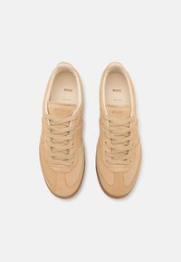 Beige suede sneakers with a low profile, rounded toe, and flat laces. Features subtle branding on the tongue and soft interior lining.