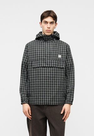 Młody mężczyzna ubrany w czarno-białą kratowaną bluzę z kapturem i elastycznymi mankietami oraz ciemne spodnie, stojący na tle jednolitego jasnego tła.