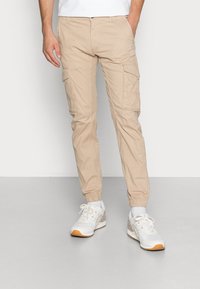 Beige cargobroek met elastische boorden, met meerdere zijzakken en een soepele katoenstructuur, gecombineerd met witte sneakers met beige accenten.