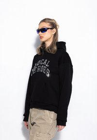 Czarna bluza z kapturem z przednią kieszenią i napisem "LOCAL HEROES" w odblaskowym druku. Model łączy ją z jasnobeżowymi spodniami cargo i okularami przeciwsłonecznymi.