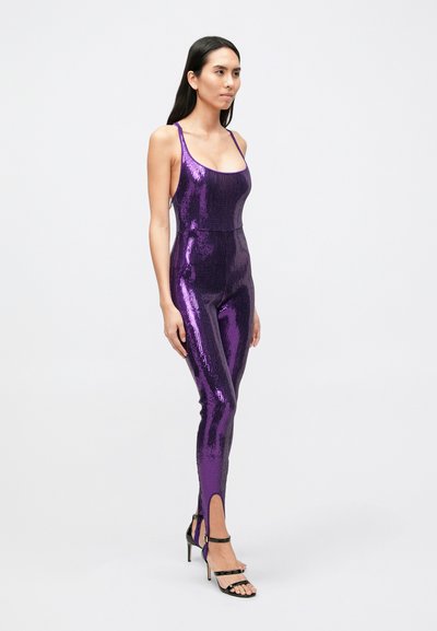 Combinaison violette à sequins avec bretelles spaghetti, silhouette ajustée, fentes latérales aux chevilles, et texture brillante. Associée à des talons noirs à lanières.
