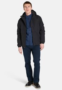 En ung man står iförd en svart huvjacka över en blå zip-up-tröja, kombinerad med marinblå byxor och svarta skor mot en enhetlig bakgrund.