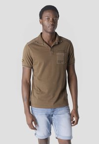 Mladi moški stoji obrnjeno naprej, oblečen v rjavo polo majico s kratkimi rokavi in svetlo modre zavihane kavbojke.