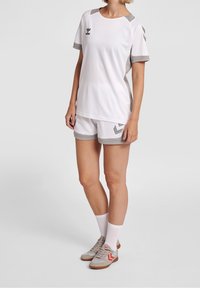 Vit sporttröja med grå detaljer och logotyp, kombinerad med matchande shorts med ett chevronmönster. Bärs med grå sneakers och vita strumpor.