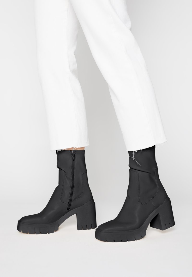 Bottes noires mi-mollet avec talons épais et fermetures éclair latérales, portées avec un pantalon blanc raccourci aux bords effilochés.