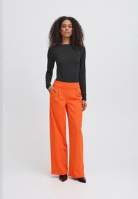 Zwarte langemouwtop en feloranje brede broek met voorzakken en een platte tailleband, voorzien van een gladde textuur en een getailleerde pasvorm.