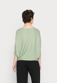 Groene gebreide top met een ruime halslijn, verlaagde schouders en een losse pasvorm. Eindigt bij de taille en is gecombineerd met zwarte broek. Zachte textuur, lichtgewicht.