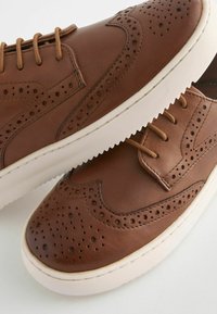 Hnědé kožené tenisky s brogue detaily, kulatou špičkou a krémovou gumovou podrážkou. Mají ploché šňůrky a perforované detaily.