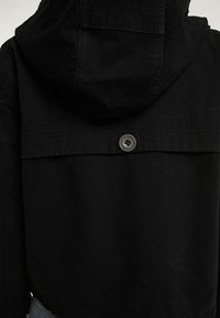 Schwarze Kapuzenjacke mit einer klaren, matten Textur, ausgestattet mit einem Rückenschluss durch Knöpfe und dezenten horizontalen Steppdetails.