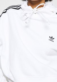 Vit huvtröja i mjukt tyg, med svart trärandig design på axlarna och en liten svärtad broderad logotyp.