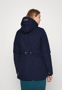 Giacca isolata di colore blu navy con cappuccio. Presenta un cordino in vita, polsini elastici e una toppa con logo vicino all'orlo.