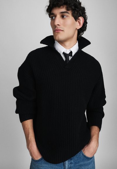 Pull en tricot noir à côtes avec un col large et des manches courtes, porté sur une chemise blanche et une cravate noire. Coupe décontractée, tissu texturé.