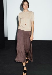 Il modello indossa un top beige e una gonna plissettata di un marrone scuro, con una collana con pendente, posando su uno sfondo minimalista.