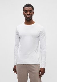 Balta ilgomis rankovėmis marškiniai, pagaminti iš minkštos medžiagos, su apvaliu iškirpimu ir įprastu siluetu. Dėvimi su šviesiai smėlinėmis kelnėmis.