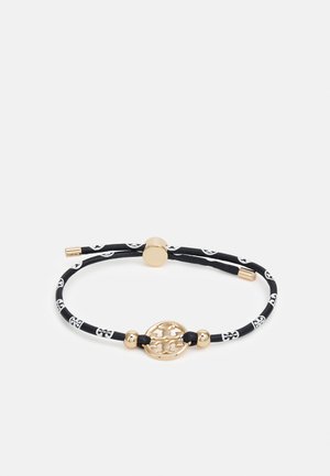 Pulsera ajustable de cordón con patrón en blanco y negro, con emblema central redondo dorado y detalles de cuentas doradas.