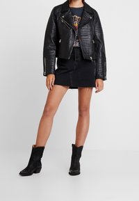 Žena v černé kožené bundě, grafickém tričku, černé denimové minisukni a černých kotníkových botách stojí proti bílému pozadí.