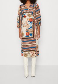 Een wikkeljurk in crème met kleurrijke bloemen- en geometrische patronen, met driekwartmouwen en een strik in de taille, gecombineerd met kniehoge witte laarzen.