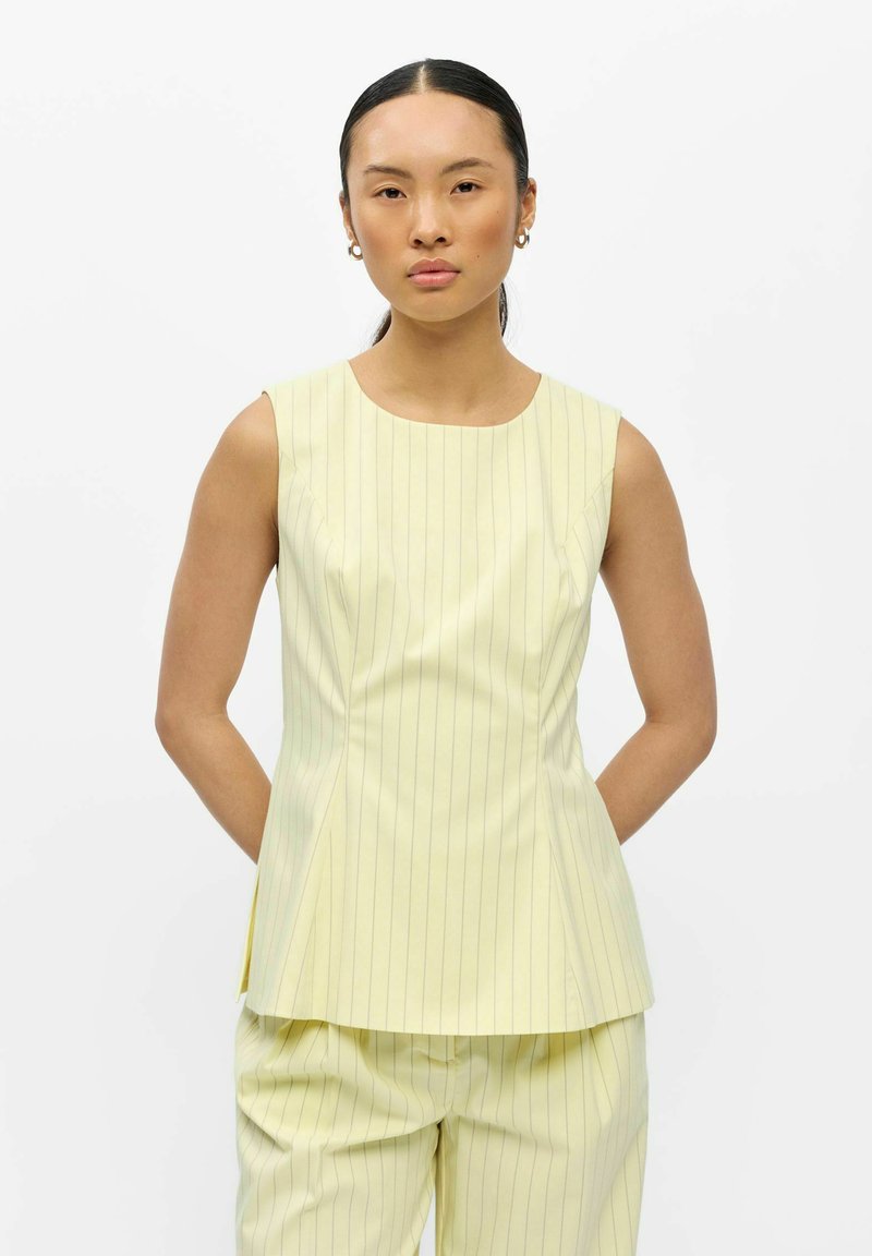 Femme aux cheveux noirs et raides portant des boucles d'oreilles créoles dorées, un haut sans manches jaune pâle à fines rayures et un pantalon ample assorti sur fond blanc.
