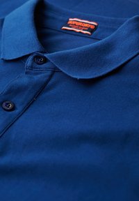 Polo shirt blu navy realizzato in tessuto testurizzato, con colletto, due bottoni e un'etichetta con il logo "Superdry" in colori a contrasto.