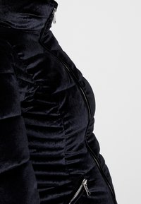 Veste de doudoune en velours noir avec un design structuré, texture matelassée verticale et zips en métal argenté sur les poches avant.