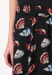 Jupe noire avec des motifs de coquillages rouges, bleus et blancs portée par une personne avec un bras gauche visible sur fond blanc.