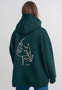Groene hoodie met een minimalistische lijntekening van een gezicht en bladeren op de achterkant, gemaakt van zachte stof met geribbelde manchetten.