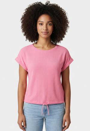 Giovane donna con capelli ricci che indossa una maglietta rosa a maniche corte con coulisse in vita e jeans azzurri, in piedi davanti a uno sfondo semplice.