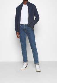 Person som bär en marinblå kabelstickad zip-up tröja, vit t-shirt, uppvikt blå jeans och vita sneakers med svarta accenter.