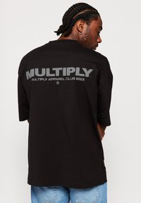 Musta ylipitkä t-paita, joka on valmistettu puuvillasta. Selässä on suuri harmaa "MULTIPLY" -logo, jonka alla lukee "MULTIPLY APPAREL CLUB 2023".