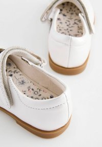 Weiße Lederschuhe mit abgerundeter Form, Klettverschluss, hellbrauner Gummisohle und floral gemustertem Innenfutter. Glatte Textur mit Nahtdetails.