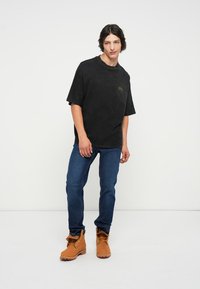 Svart t-skjorte med korte ermer som har en subtil grafikk, kombinert med blå denimjeans og brune støvler, som viser en uformell, avslappet passform.