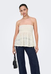 Weißes, trägerloses Peplum-Top aus strukturiertem Stoff mit offenen Stickmustern. Kombiniert mit blauen weitem Jeans und einer kleinen schwarzen Handtasche.