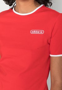 Červená košile s krátkým rukávem a kulatým výstřihem, s bílými lemy a vyšitým logem Adidas v bílé a červené barvě na levé hrudi.