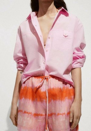 Osoba ubrana w luźną różową koszulę na guziki z podwiniętymi rękawami oraz różowo-pomarańczowe szorty tie-dye z wiązanym paskiem w talii na jednolitym tle.