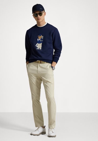 Sweatshirt marine avec graphique d'ours brodé, pantalons beiges, chaussures blanches et une casquette marine assortie. Des lunettes de soleil complètent le look.
