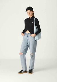 Jovem mulher vestindo uma blusa preta com fecho, jeans claros rasgados, boné preto, tênis pretos, segurando uma bolsa de ombro azul clara, de pé contra um fundo branco.
