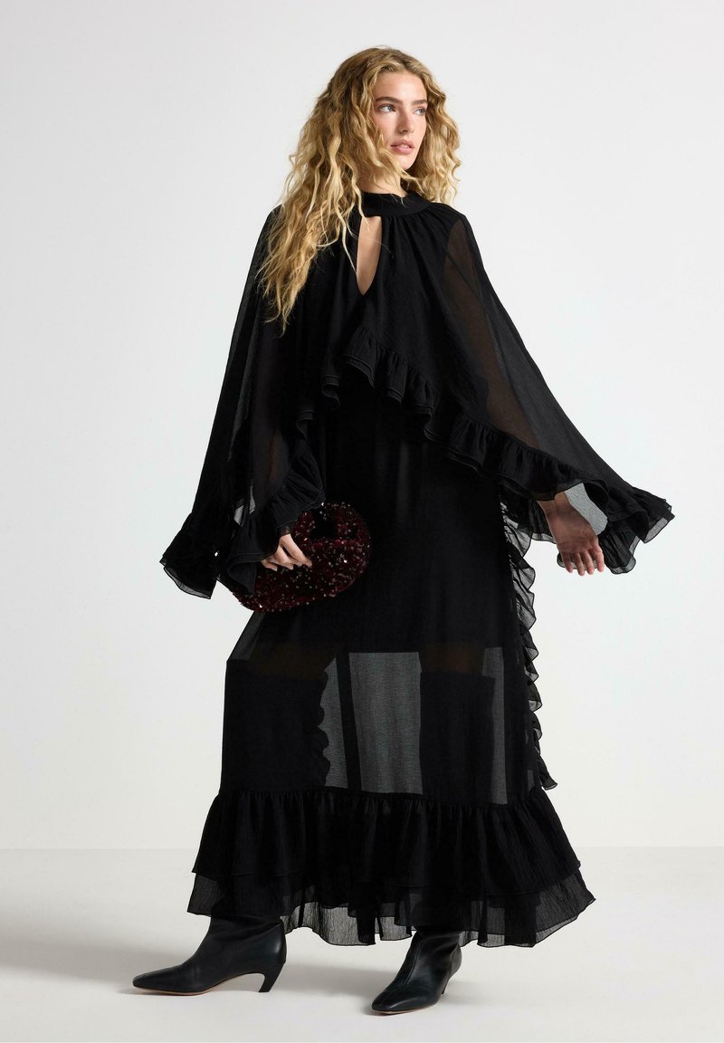 Svart genomskinlig maxiklänning med volanger, hög halsringning och nyckelhålsutskärning. Matchad med en strukturerad vinröd clutch och svarta spetsiga stövlar.