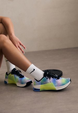 Ben iført hvide Nike-sokker og multifarvede Nike-sports sko, hvilende på en glat gulv nær en vægtplade.