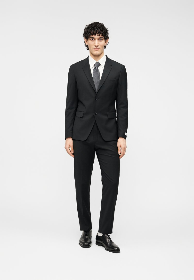 Μαύρο κοστούμι με σακάκι εφαρμοστό, ταιριαστό παντελόνι και λευκό πουκάμισο. Γκρι γραβάτα και μαύρα γυαλισμένα παπούτσια ολοκληρώνουν την εμφάνιση.