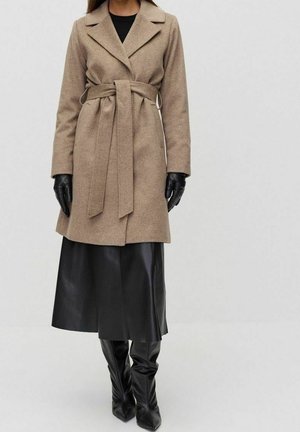 Mujer con un abrigo beige con cinturón hasta la rodilla, guantes de cuero negro, falda midi negra y botas negras hasta la rodilla, posando sobre un fondo liso.