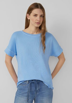 Young woman with long light brown hair wearing a loose light blue short-sleeve top and blue jeans, standing against a plain background.