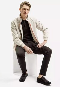 Jasno brązowa kurtka z czerwonymi akcentami, czarna koszulka polo, ciemne dopasowane spodnie i czarne sneakersy. Gładkie faktury, swobodny design.