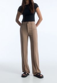 Beige hoge taille broeken met voorzakken, zichtbare naden en een enkele knoopsluiting, gecombineerd met een aansluitend zwart topje en zwarte sandalen.