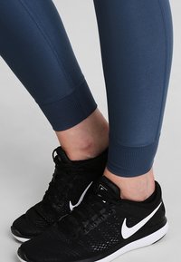 Leggings bleu marine en tissu lisse et extensible avec des poignets côtelés, associés à des chaussures de sport noires avec un détail en swoosh blanc.