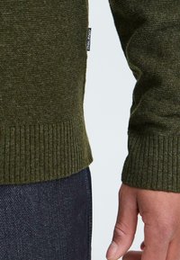 Olijfgroene gebreide trui met geribbelde manchetten en zoom, met een zachte textuur. Een klein label aan de zijkant zegt "BLEND." Donkere spijkerbroek is gedeeltelijk zichtbaar.