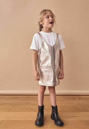Rochie metalizată argintie cu două buzunare frontale, suprapusă peste un tricou alb, combinată cu botine negre până la gleznă, pe un fundal bej.