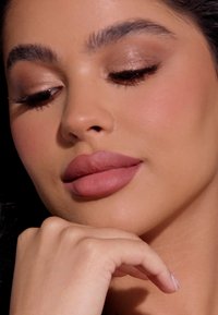 Close-up van een gezicht met een gladde, gebruinde huid, volle matte mauve lippen, zachte oogmake-up met subtiele glans en donkere wimpers. Een vinger rust op de kin.