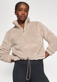 Pull en polaire beige avec un zip quart, un col haut, un ourlet et des poignets élastiques, doté d'un cordon de serrage à la taille et avec une texture douce.
