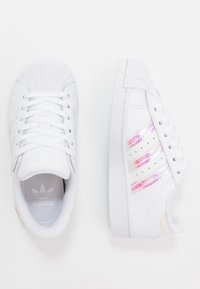 Vita sneakers med texturerad tå, skosnören och tre iriserande ränder på sidorna. Utrustade med en slät, gummisula och vadderad insida.